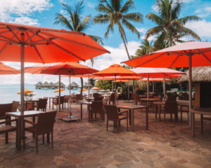 outdoor restaurant with orange umbrellas and lots of square tables in bora bora at the intercontinental bora bora le moana resort with overwater bungalows and palm trees in the backdrop