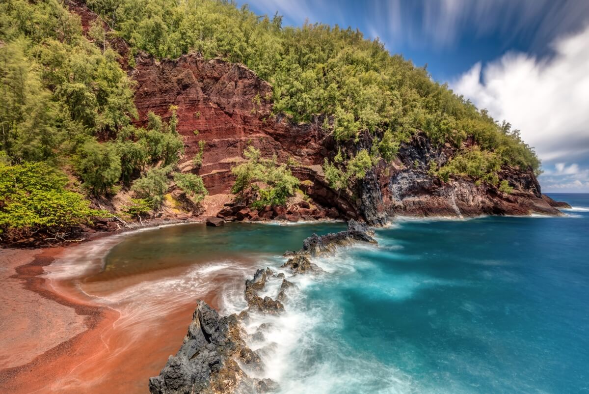 red sand beach with waves crashing along it surrounded by greenery and rocks on the road to hana in maui hawaii