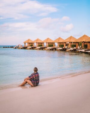 woman with colorful flowing top on sitting on the edge of water on the beach in front of overwater cabanas at renaissance private island in aruba