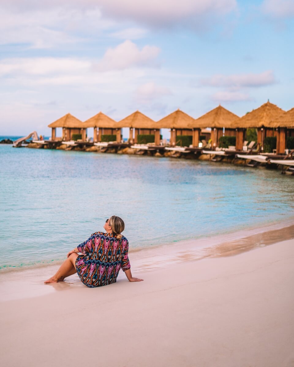 woman posing while sitting on beach in front of overwater cabanas in aruba