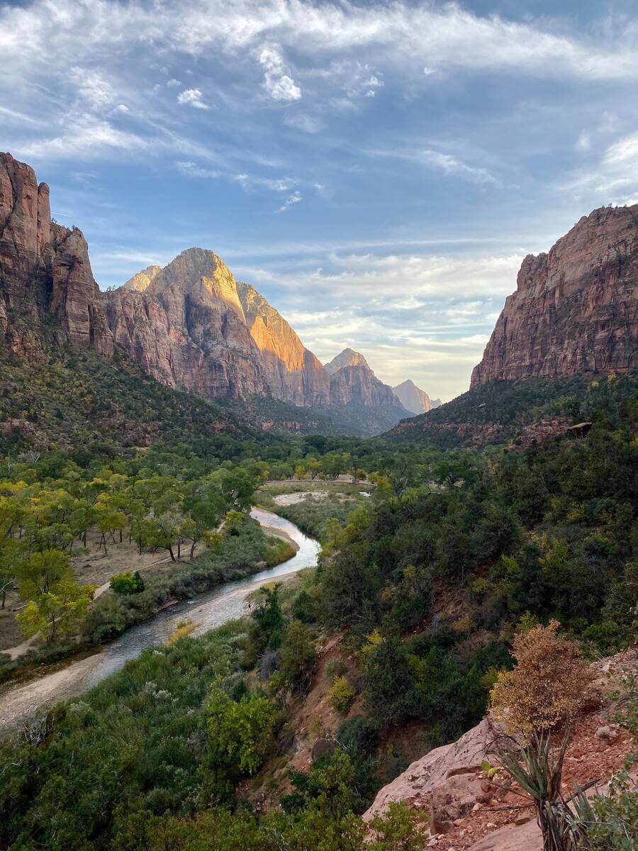 valley in zion national park with a river running through the frame and green trees surrounding on either side with mountains in the distance