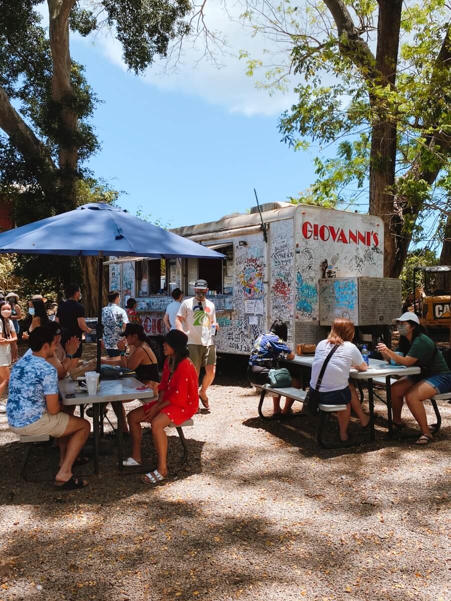 picnic tables full of people in shaded area with giovanni's shrimp food truck in the background, which is a white truck covered in writing an graffiti in oahu