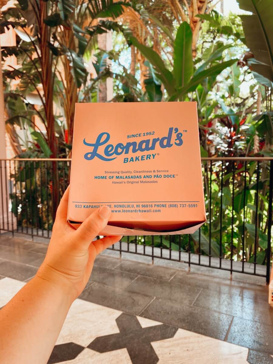 woman's hand holding pink leonard's bakery box of malasadas, which are fried portguese donuts, in front of tropical and lush hawaiian backdrop