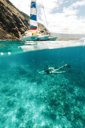 woman and man snorkeling in the ocean underneath colorful catamaran boat, things to do big island hawaii
