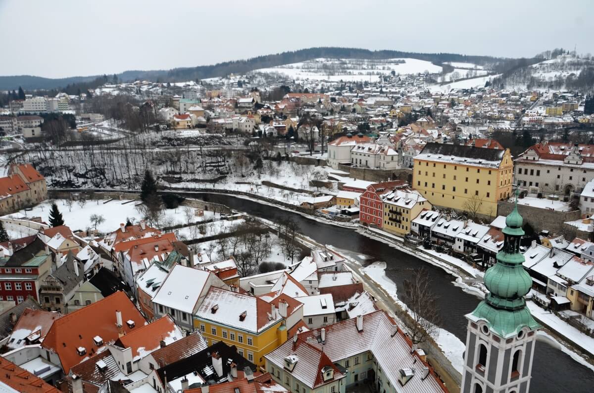 snow covered view of the city of cesky krumlov, with orange roofs and a small river going throughout the city