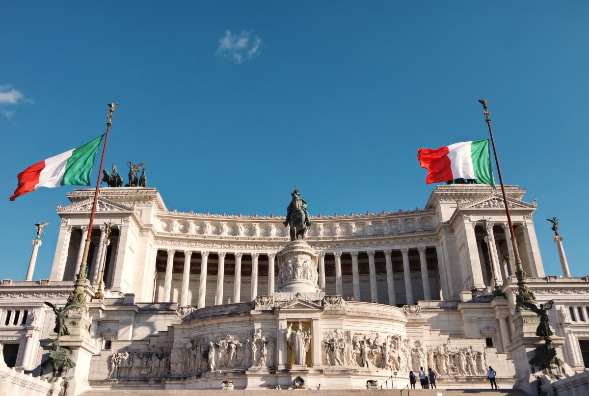 stunning building and statues with italian flags hanging on flag posts below a blue sky in rome