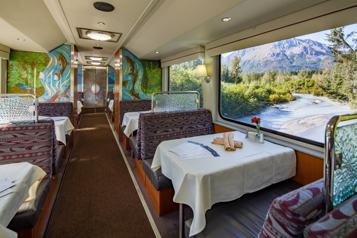 photo of dining car on alaska railroad with tables set with white table clothes and cutlery, with the alaskan wilderness in the background out the window