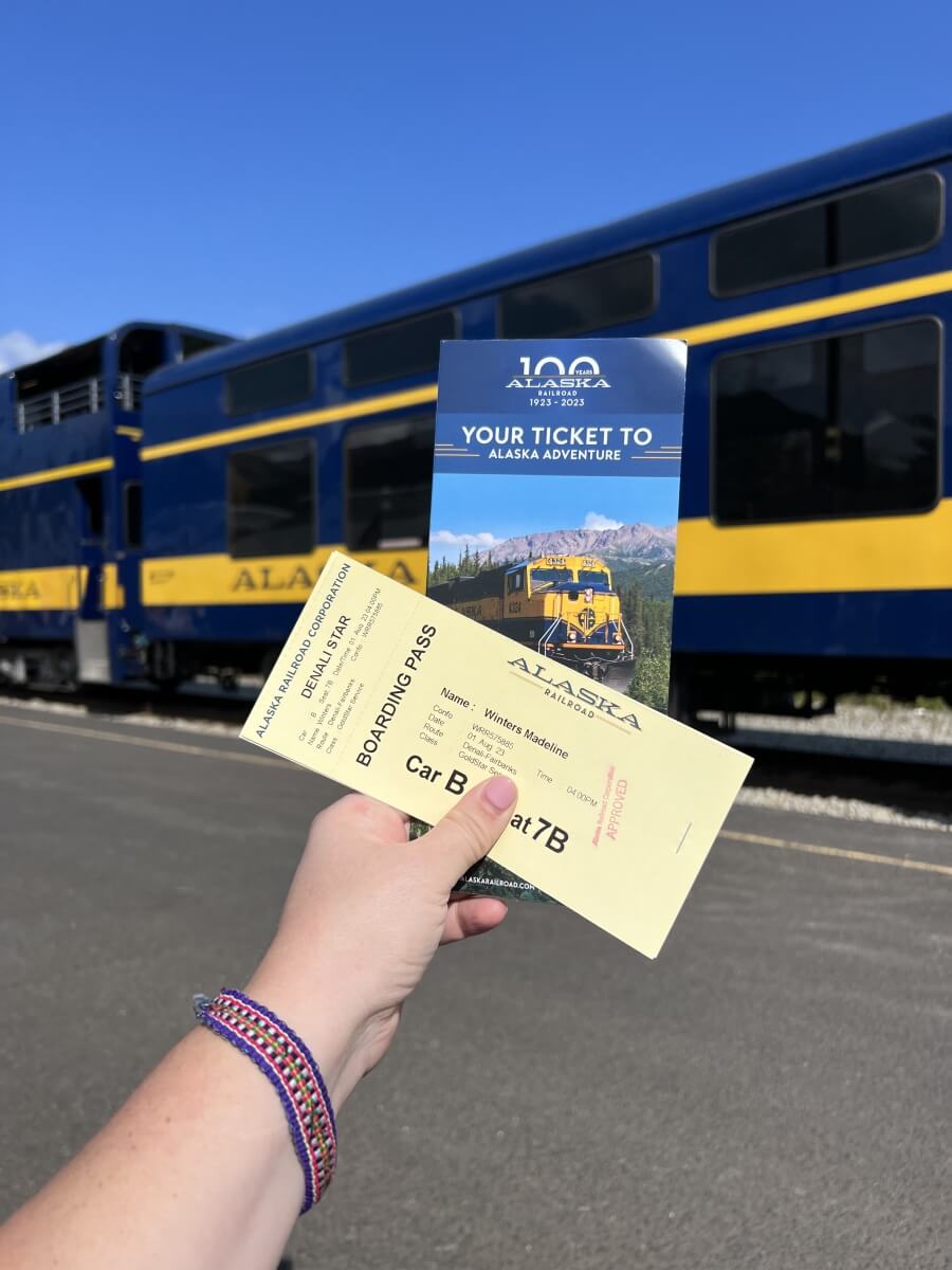 woman holding alaska railroad yellow ticket stub with colorful pamphlet detailing alaska railroad in front of alaska railroad train on a sunny day with blue skies