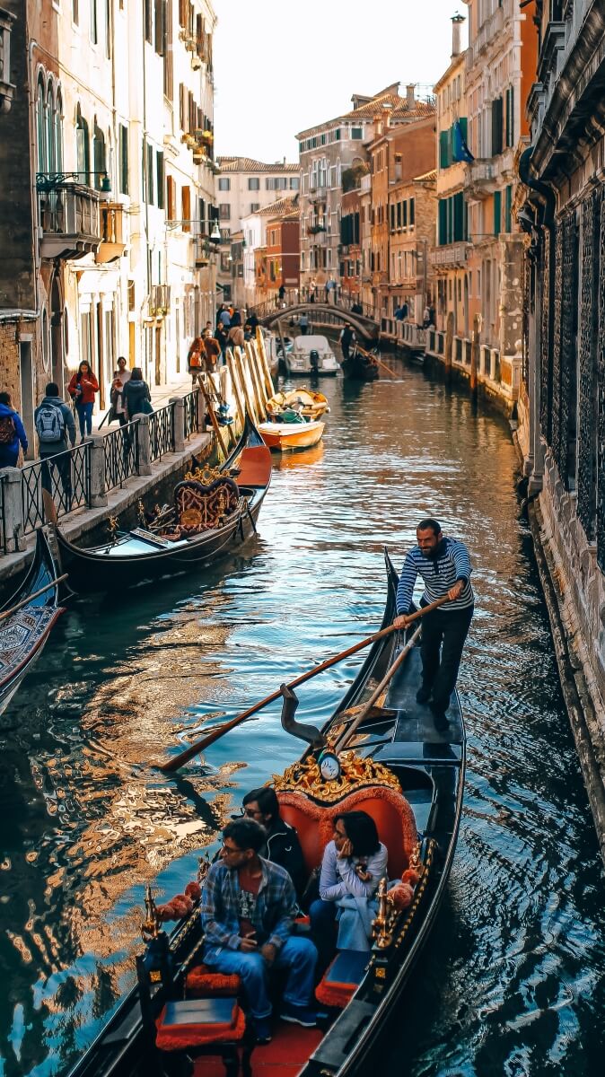 man rows gondola down the canals in venice italy with other gondolas lining the side of the canals as people walk by historic buildings, one of the best things to do in Venice Italy