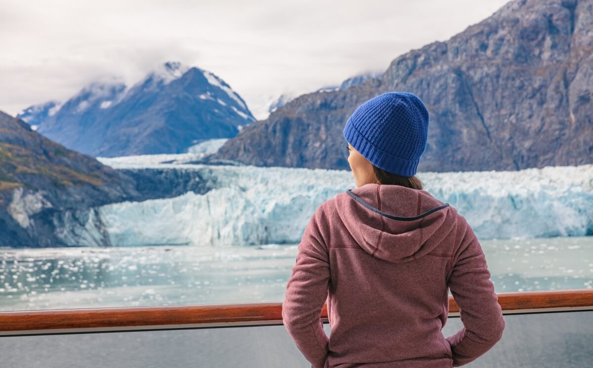 woman with pink jacket and blue beanie hat looking over alaska cruise balcony cabin to glaciers in alaska with mountains all around