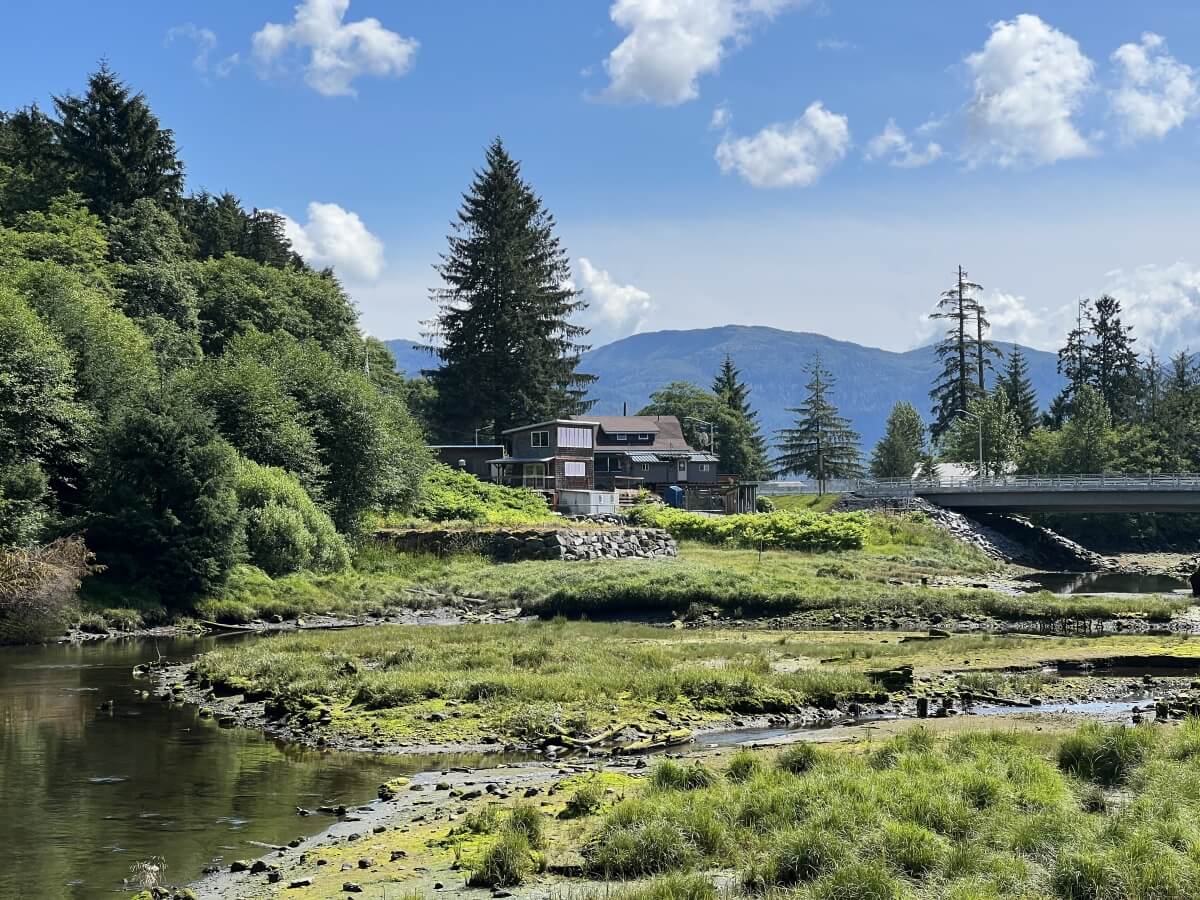 river bank area in alaska surrounded by greenery, blue skies, and mountains with gray home in the center frame | alaska cruise booking tips