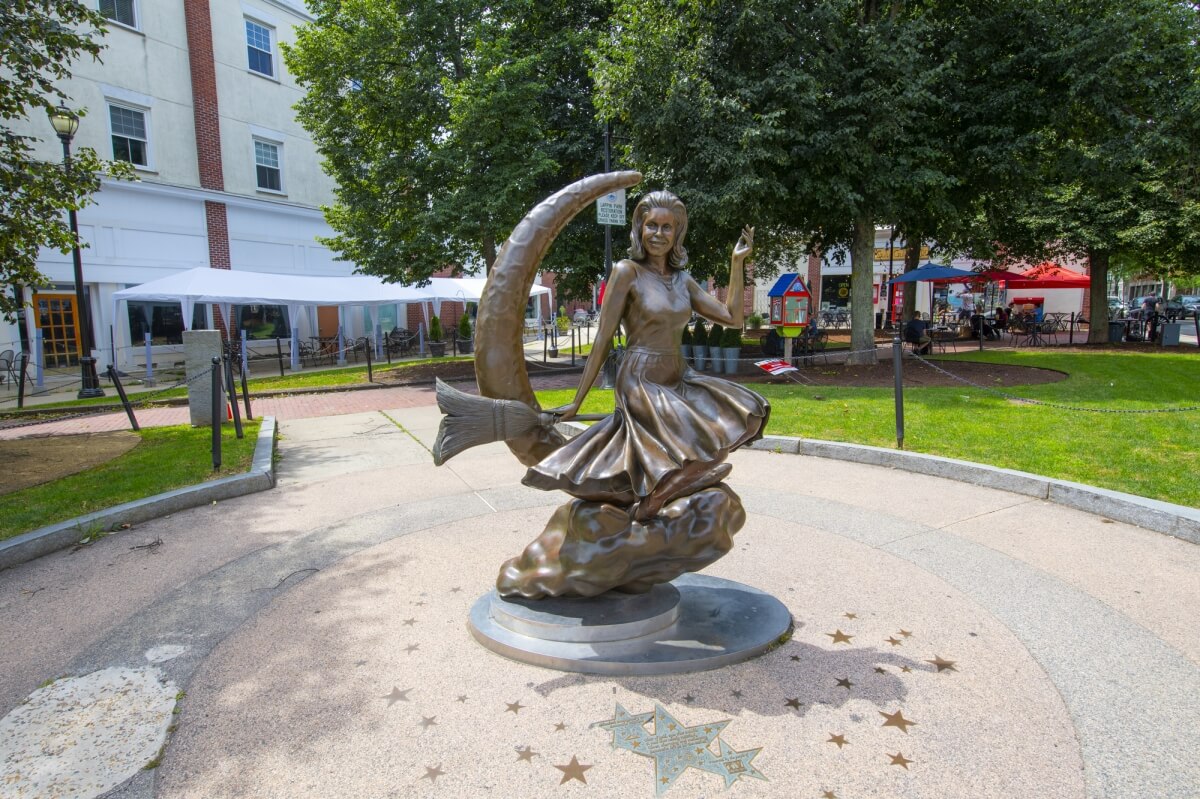 the bewitched statue in salem massachussetts - a bronze statue with a woman in a dress sitting on a broomstick in front of a crescent moon in a city square