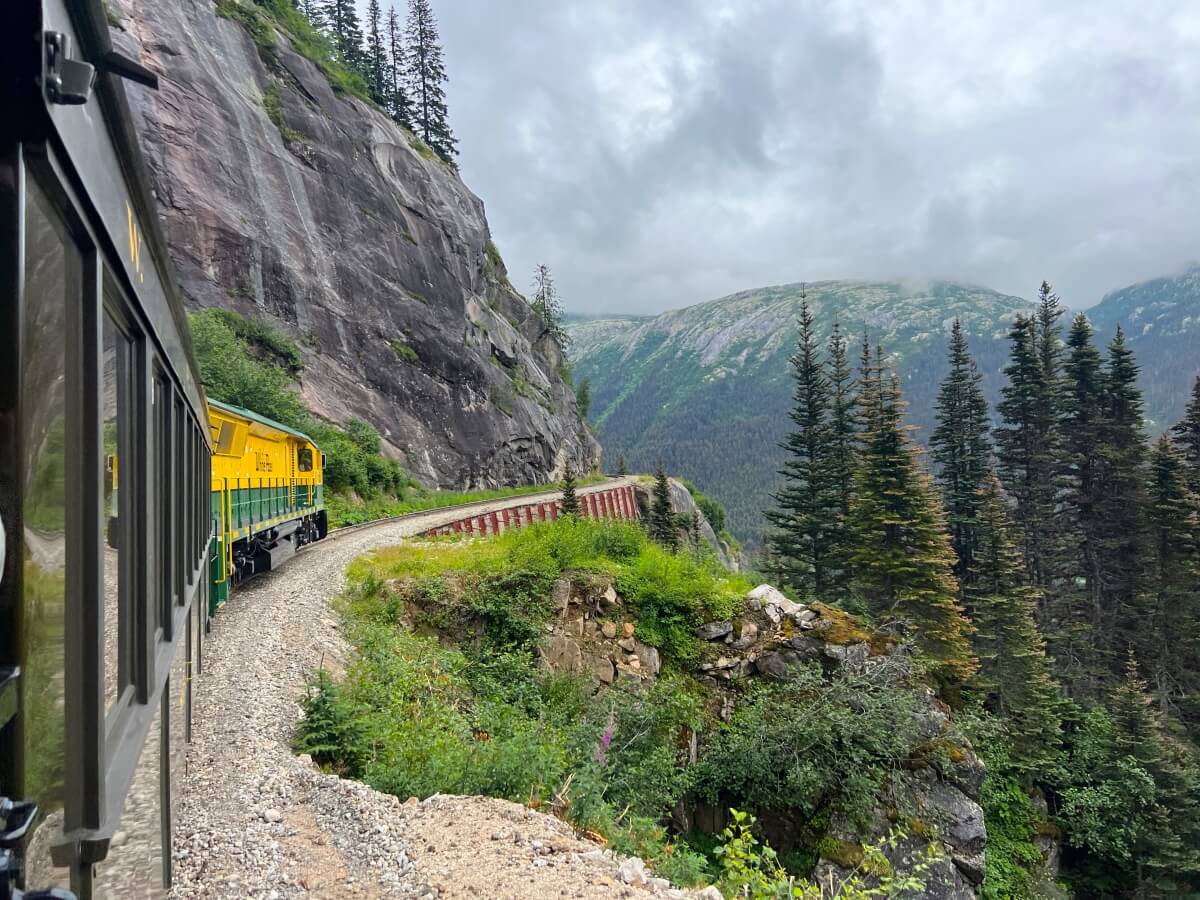 yellow and green train on tracks along the side of a mountain, surrounded by lots of trees and other greenery