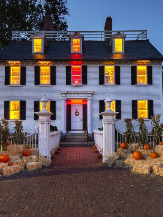 a white house with windows lit up in yellow and red lights with hay stacks and pumpkins out front of the white fence