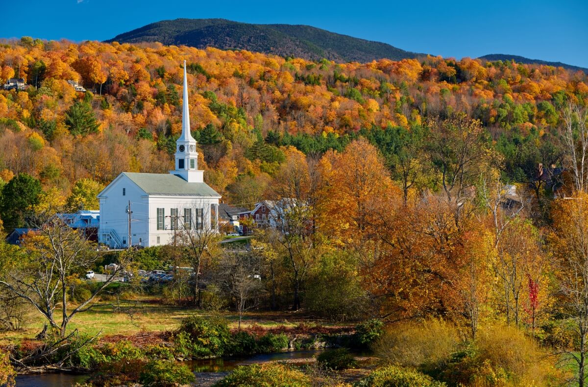 a white church with a large steeple sitting amongst green, orange, yellow, and red trees, in front of a mountain in the backdrop