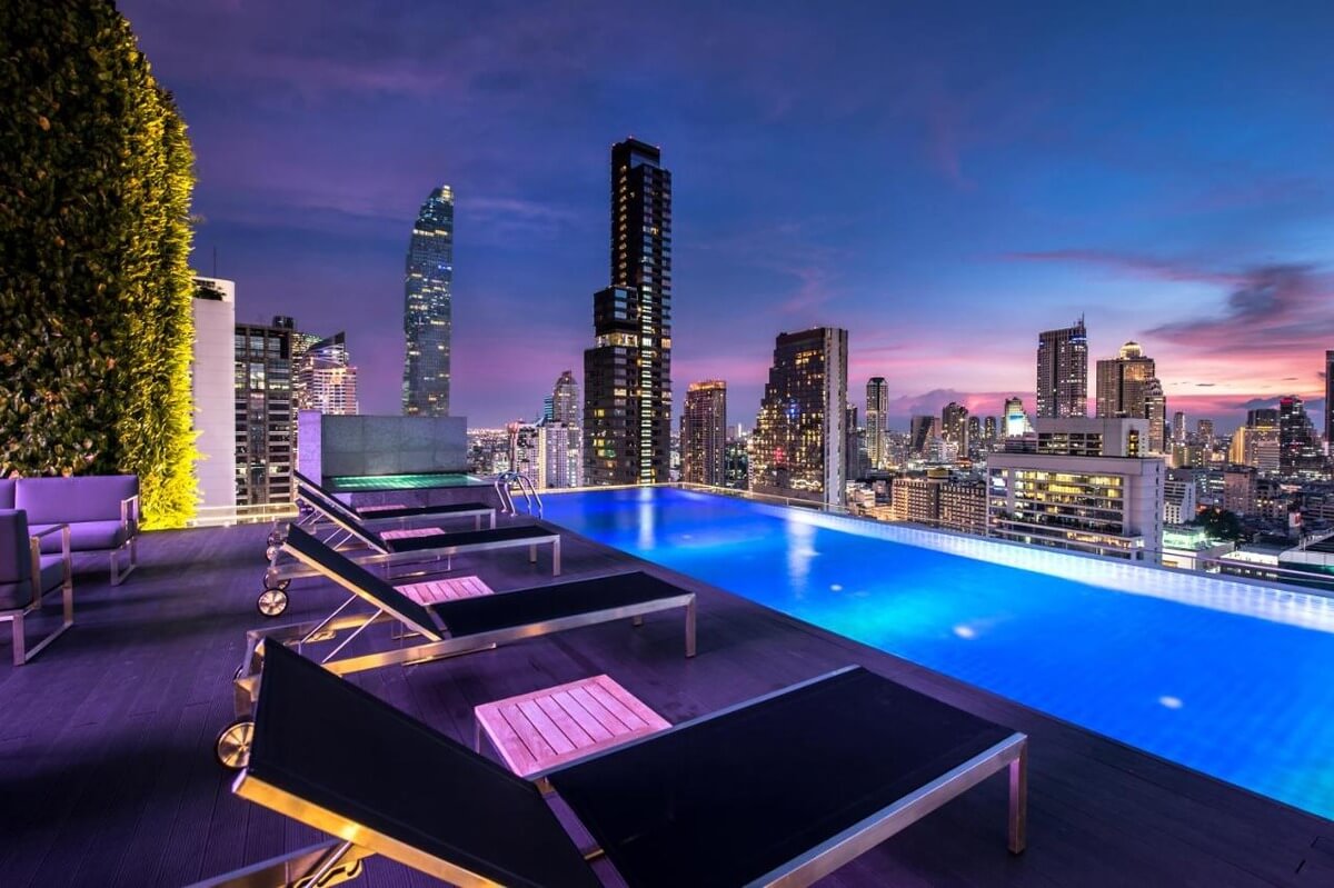 The infinity pool at the Amara hotel in Bangkok at night lit up by blues and purples and the sun setting in the distance